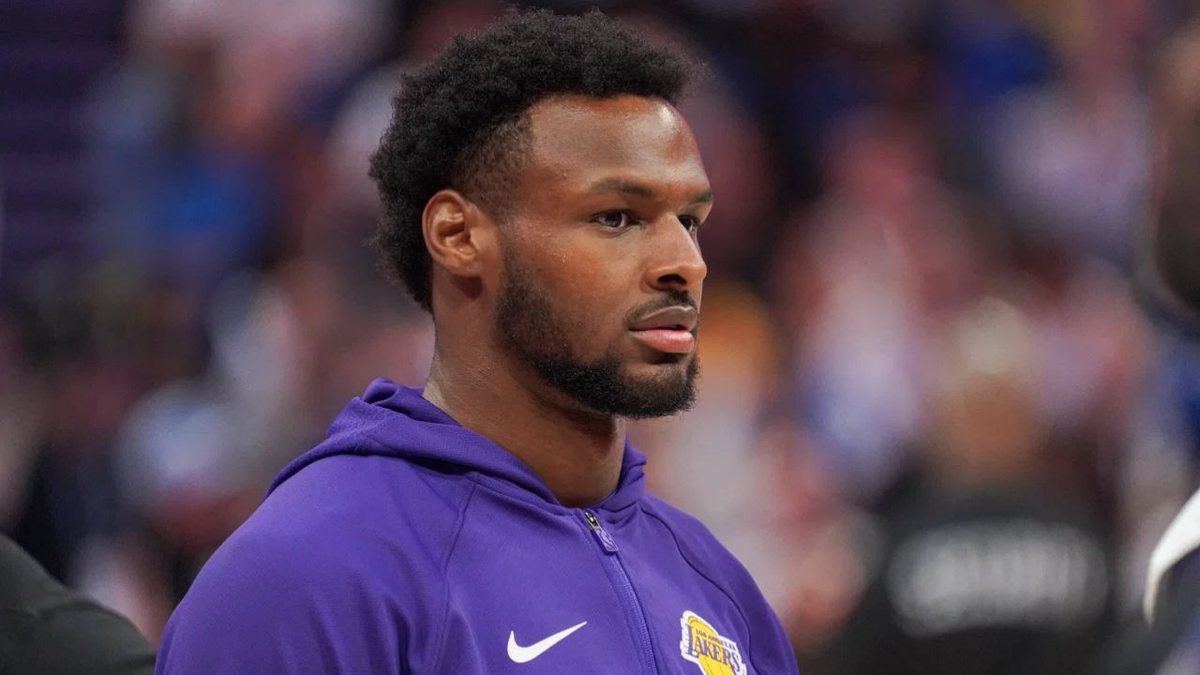 Bronny James, in a purple Lakers hooded warmup jacket, stands on the floor before a game.