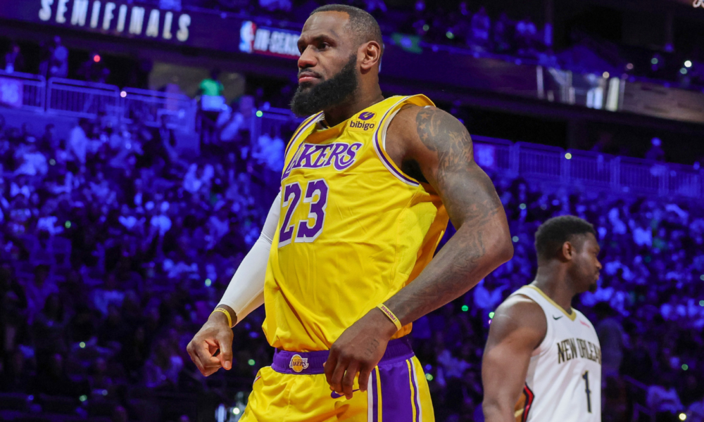 LeBron James wearing a yellow Los Angeles Lakers jersey, standing on the court during a game against the New Orleans Pelicans, with a focused expression and the crowd illuminated in blue and purple tones in the background.