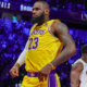 LeBron James wearing a yellow Los Angeles Lakers jersey, standing on the court during a game against the New Orleans Pelicans, with a focused expression and the crowd illuminated in blue and purple tones in the background.