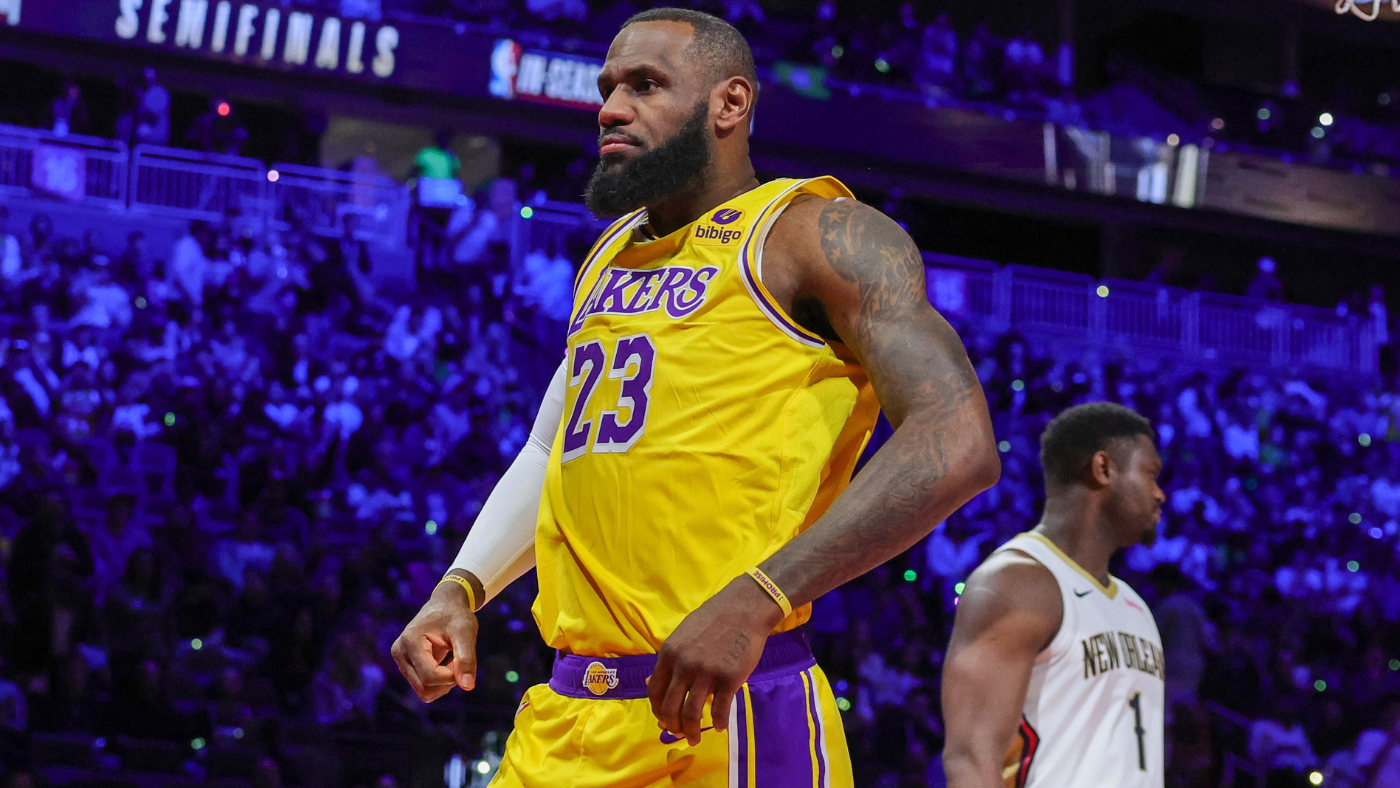 LeBron James wearing a yellow Los Angeles Lakers jersey, standing on the court during a game against the New Orleans Pelicans, with a focused expression and the crowd illuminated in blue and purple tones in the background.